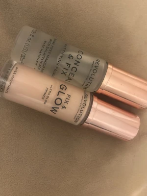 Revolution Fix & Glow och Conceal & Fix Primer - Två primers från Revolution: Fix & Glow Ultra Glow Primer i ljus beige nyans med roséguldfärgat lock och Conceal & Fix Mattifying Primer i transparent formula med roséguldfärgat lock. Båda i glasflaska, perfekta för att skapa en jämn bas.