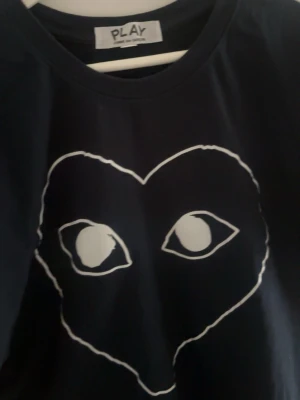 Svart PLAY t-shirt med hjärtmotiv - Snygg svart t-shirt från Comme des Garçons PLAY med det ikoniska vita hjärtmotivet och ögon på bröstet. Klassisk rund hals och korta ärmar. Perfekt för dig som gillar streetwear och vill sticka ut med en unik design. Är liten i storleken så skulle säga att den passar mera än L än XL. Ge gärna bud.