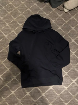 Marinblå hoodie med broderad logga - Snygg svart hoodie  med broderad logga på bröstet. Klassisk modell med huva och dragsko, tillverkad i mjukt material som känns skönt mot huden. Perfekt för en stilren och avslappnad look.