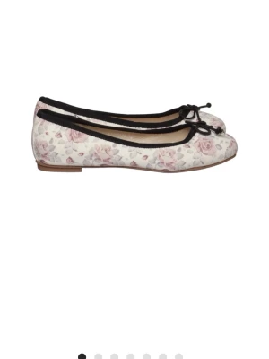 Blommiga ballerinaskor med rosett - Söta ballerinaskor med vit bas och ljusrosa blommönster. Skorna har svarta kanter och en liten rosett framtill. Platt sula och rund tå ger en klassisk look. Perfekta för dig som gillar romantisk och feminin stil.