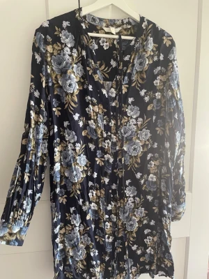 Blommig långärmad klänning H&M  - Mörkblå långärmad klänning från H&M med vackert blommigt mönster i blått, beige och vitt. Klänningen har knytband, bekväm passform. Perfekt för dig som vill ha en stilren och avslappnad look.