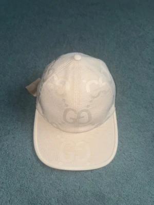 Vit keps från Gucci med GG-mönster - Snygg vit keps från Gucci med klassiskt GG-monogram över hela kepsen. Kepsen har böjd skärm, justerbar rem baktill och är tillverkad i canvasmaterial. Perfekt för dig som vill ha en stilren och lyxig accessoar till din outfit.
