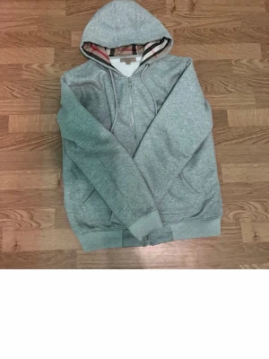 Grå hoodie Burberry med dragkedja - Snygg grå hoodie från Burberry med dragkedja framtill och klassiskt rutigt foder i huvan. Hoodien har fickor på sidorna och diskret Burberry-logga broderad på bröstet. Hör av dig för mer info!