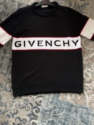 Svart stickad tröja från Givenchy - Cool svart stickad tröja från Givenchy med vit rand och logotyp över bröstet. Ärmarna har vita ränder med röda detaljer. Tröjan har rund halsringning och ribbade muddar. Perfekt för dig som vill ha en trendig och exklusiv look.