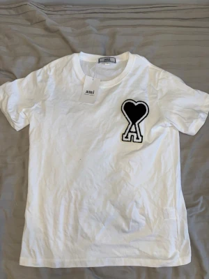 Vit t-shirt från Ami Paris - Vit t-shirt från Ami med svart hjärta och A-logga på bröstet. Klassisk rund halsringning och korta ärmar. Tillverkad i mjuk bomull för en skön känsla. Perfekt för en clean och stilren look.