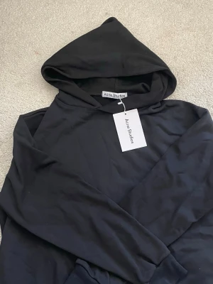 Svart hoodie från Acne Studios - Snygg svart hoodie från Acne Studios med klassisk passform och huva. Tröjan har ribbade muddar vid ärmslut och nederkant samt en stilren look utan tryck. Perfekt för dig som gillar minimalistisk och trendig streetwear.