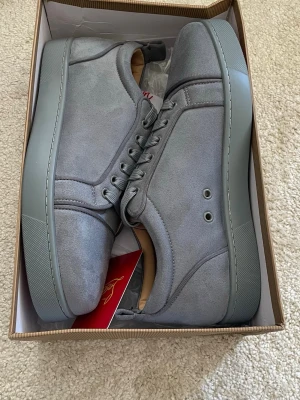 Gråa sneakers från Christian Louboutin - Stilrena gråa sneakers från Christian Louboutin i mocka med matchande grå sula och snörning. Skorna har diskreta detaljer och en klassisk låg siluett. Insidan är fodrad i beige läder för extra komfort. Perfekta för dig som gillar minimalistisk och lyxig stil.