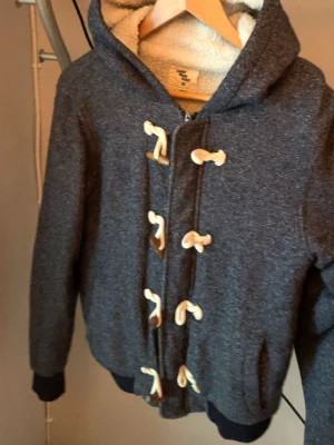 Mörkgrå teddyjacka med snörning - Mysig mörkgrå teddyjacka med huva och vitt fluffigt foder. Jackan har dekorativa snörningar framtill och ribbade muddar vid ärmar och nederkant. Perfekt för kyliga dagar och ger en avslappnad look.