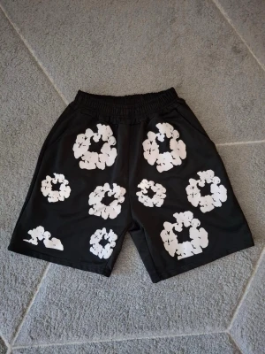 Svarta shorts med vita blommor Denim Tears - Svarta shorts från Denim Tears med elastisk midja och stora vita blommönster över hela plagget. Perfekta för en avslappnad och trendig stil. Materialet känns mjukt och bekvämt, och designen sticker verkligen ut.