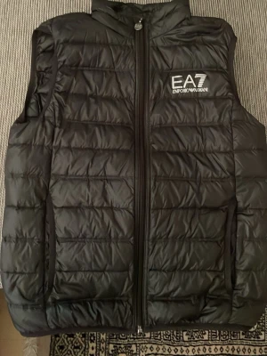 Svart dunväst från Emporio Armani EA7 - Stilren svart dunväst från Emporio Armani EA7 med quiltad design och hög krage. Västen har dragkedja framtill och en vit logotyp på bröstet. Perfekt för lager-på-lager och ger en sportig look. Inga hål eller skador. Bara att höra av sig vid frågor👍🏼
