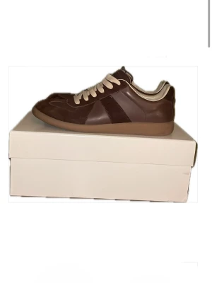 Bruna Replica sneakers från Maison Margiela -  Priset kan diskuteras och kan skicka fler bilder Snygga bruna Replica sneakers från Maison Margiela med beige mockadetaljer och vita snören. Skorna har en klassisk låg silhuett och gummisula. Insidan är vit och märkt med Maison Margiela-loggan. Perfekta för dig som gillar stilrena och tidlösa sneakers.