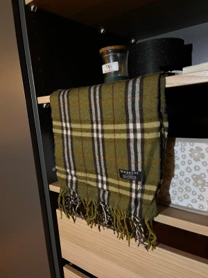 Grön rutig halsduk från Burberry - Säljer en Snygg grön halsduk från Burberry i klassiskt rutigt mönster med inslag av vitt, svart och brunt. Tillverkad i 100% lammull och har fransar i ändarna. Perfekt accessoar för att lyfta din outfit med en tidlös och lyxig känsla.
