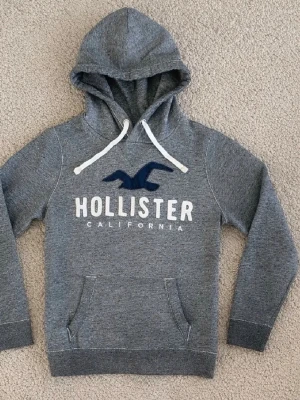 Grå hoodie från Hollister - Mysig grå hoodie från Hollister med stor logga och texten 'Hollister California' på bröstet. Hoodien har vit dragsko i huvan och en klassisk känguruficka framtill. Tillverkad i mjukt sweatshirtmaterial och har ribbade muddar vid ärmar och nederkant.