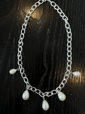 Silverfärgat halsband med pärldroppar - Stilrent halsband i silverfärgad metall med stora länkar och fyra droppformade vita pärlor som hängsmycken. Perfekt för att ge en cool touch till din outfit. Kedjan har en chunky känsla och pärlorna ger en elegant kontrast.
