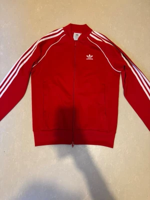 Röd Adidas track jacket - Klassisk röd Adidas track jacket med vita ränder längs ärmarna och vit Adidas-logga på bröstet. Dragkedja framtill, ribbade muddar och två fickor. Tillverkad i ett mjukt och stretchigt material som ger en sportig look.
