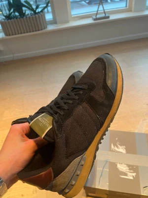 Valentino Garavani svarta sneakers - Säljer ett par svarta Valentino Garavani sneakers med coola nitar på hälen, bruna läderdetaljer och beige sula. Ovandelen är i mesh och mocka för en schysst mix av material. Skorna har klassisk låg profil och snörning framtill. Liten slitning vid hällen men inget hål! Är i storlek 45 men har själv 44 och passar perfekt!