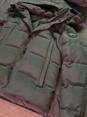 Grön dunjacka från Canada Goose - Säljer en grön dunjacka från Canada Goose med klassisk patch på ärmen. Jackan har huva, dragkedja och tryckknappar framtill samt två stora fickor. Fylld med dun för extra värme och perfekt för kalla vinterdagar. Snygg och tidlös design som håller dig varm.