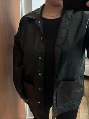 Svart regnjacka med knappar - Snygg svart jacka i glansigt material med tryckknappar framtill och två stora fickor. Jackan har en rak passform och klassisk krage, perfekt för vardagen. Enkel och stilren design som passar till det mesta.