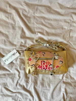 Guldig Betty Boop handväska - Snygg guldig handväska med Betty Boop-tryck och flera dekorativa berlocker i hjärt- och stjärnform. Väskan har nitar, dragkedja framtill och detaljer i rosa och rött. Axelremmen är justerbar och väskan har en unik, lekfull design med metallic-finish.
