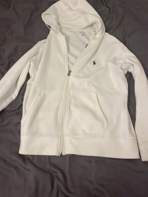 Vit hoodie från Polo Ralph Lauren - Snygg vit hoodie med dragkedja från Polo Ralph Lauren. Klassisk design med huva, två fickor framtill och den ikoniska lilla loggan broderad på bröstet. Perfekt för en clean och stilren look. Materialet känns mjukt och bekvämt.