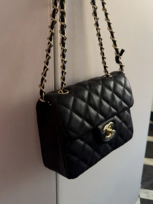 Svart Chanel axelväska i läder - Säljer en klassisk svart Chanel axelväska i quiltat läder med guldfärgade detaljer och kedjerem. Väskan har den ikoniska CC-loggan på locket och en tidlös, elegant design. Perfekt för dig som vill ha en lyxig accessoar med exklusiv känsla.