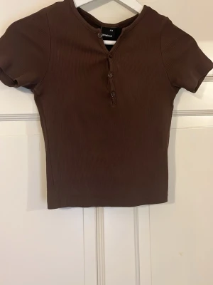 Brun ribbad topp med knappar - Säljer en brun ribbad topp från gina tricot med korta ärmar och tre knappar framtill. Toppen har en rund halsringning och är i en mjuk, stretchig bomullsblandning. Perfekt till jeans eller kjol för en enkel och snygg look.