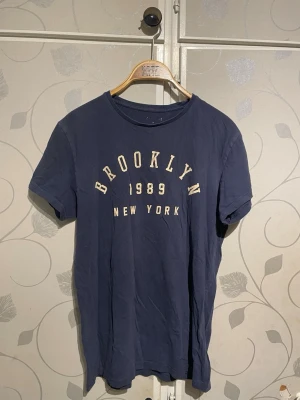 Mörkblå Brooklyn t-shirt - Snygg mörkblå t-shirt med texten 'BROOKLYN 1989 NEW YORK' i vitt och gult framtill. Klassisk rund halsringning och korta ärmar. Perfekt till jeans eller shorts för en avslappnad streetstyle-look.