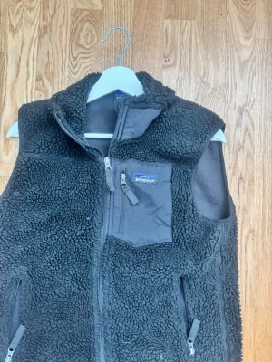 Svart  från Patagonia XS - Mysig svart teddyväst från Patagonia i storlek XS. Västen har hög krage, dragkedja framtill, tre fickor med dragkedja och detaljer i nylon på bröstfickan. Perfekt för lager-på-lager och riktigt skön att bära under kyliga dagar.