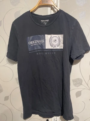 Svart t-shirt från Jack & Jones - Snygg svart t-shirt från Jack & Jones med tryck framtill där det står 'ORIGINALS' och 'WORLDWIDE'. T-shirten har rund halsringning och korta ärmar. Materialet är mjuk bomull som känns skönt mot huden. Perfekt till jeans eller shorts.