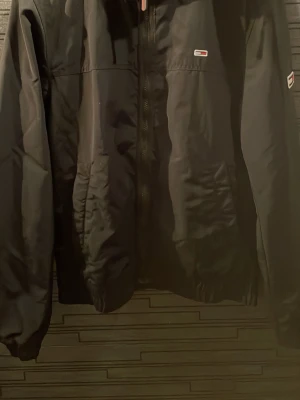 Svart vindjacka från Tommy Hilfiger - Stilren svart vindjacka från Tommy Hilfiger med dragkedja framtill och små logodetaljer på bröstet och ärmen. Jackan har elastiska muddar och nederkant för en skön passform. Perfekt för blåsiga dagar och enkel att matcha med olika outfits.