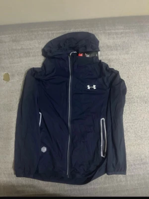 Marinblå vindjacka från Under Armour - Snygg marinblå vindjacka från Under Armour med huva och hel dragkedja framtill. Jackan har vita detaljer, två fickor med dragkedja och Under Armour-logga på bröstet. Tillverkad i lätt och funktionellt material som passar perfekt för blåsiga dagar.