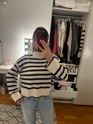 Randig stickad tröja med polokrage - Säljer en vit stickad tröja med svarta horisontella ränder och polokrage. Tröjan har en boxy, croppad passform och långa ärmar med breda ribbade muddar. Perfekt för dig som gillar en avslappnad och trendig stil.