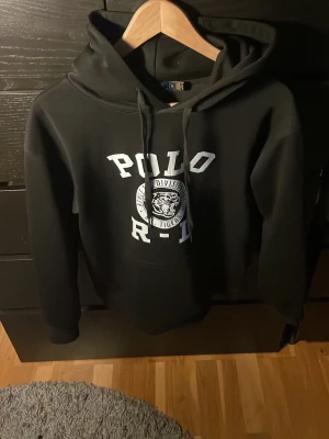 Svart hoodie från Polo Ralph Lauren - Svart hoodie från Polo Ralph Lauren med stor vit logga och text på bröstet. Klassisk huva med dragsko och mjukt material som känns skönt mot huden. Perfekt för en avslappnad och stilren look.