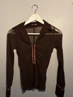 Brun meshblus med dragkedja - Trendig brun meshblus med lång ärm och markerad krage. Blusen har en dekorativ dragkedja framtill med rosa detaljer och kontrastsömmar. Materialet är genomskinligt mesh som ger en cool och modern look. 