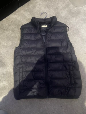 Mörkblå dunväst med dragkedja - Snygg svart dunväst med quiltad design och hög krage. Västen har dragkedja framtill och två sidofickor. Perfekt för lager-på-lager och ger en clean look till din outfit. Passar till både streetwear och casual stil.