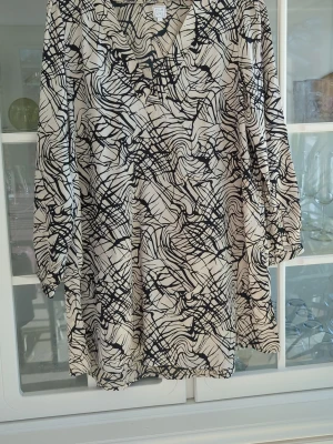 Mönstrad blus med v-ringning - Snygg blus i vitt med svart abstrakt mönster och v-ringning. Blusen har trekvartsärm med lätt puff och är tillverkad i ett mjukt, tunt tyg som faller fint. Perfekt för dig som gillar unika prints och avslappnad passform.