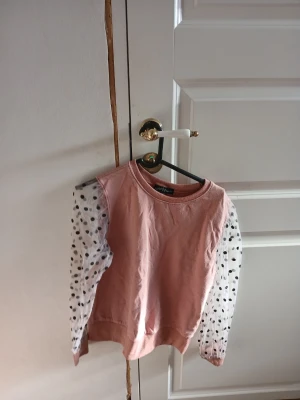 Rosa tröja med prickiga ärmar - Säljer en söt rosa tröja från KappAhl med transparenta, vita ärmar dekorerade med svarta prickar. Tröjan har rund halsringning och ribbade muddar i nederkant. Perfekt för dig som gillar unika detaljer och en lekfull stil.