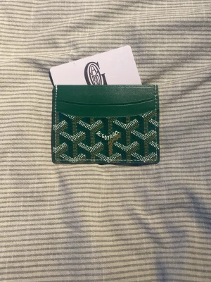 Grön  korthållare i läder - Snygg grön korthållare  med ikoniska mönster i vitt och svart. Tillverkad i läder och har flera fack för kort. Kommer med originalask, tygpåse och äkthetskort. Perfekt för dig som vill ha något exklusivt och stilrent i fickan.