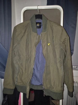 Olivgrön bomberjacka från Lyle & Scott - Snygg olivgrön bomberjacka från Lyle & Scott med klassisk gul logga på bröstet. Jackan har ribbade muddar vid ärmslut, krage och nederkant samt två fickor med dragkedja. Perfekt för dig som vill ha en stilren och trendig look.