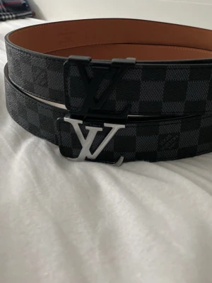 Louis Vuitton rutiga bälten - Två snygga Louis Vuitton-bälten med klassiskt rutigt mönster i svart och grått. Ena bältet har ett svart LV-spänne och det andra ett silverfärgat LV-spänne. Perfekta accessoarer för att lyfta din outfit.