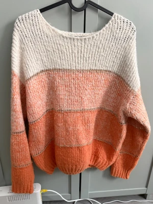 Randig stickad tröja i orange och vit - Mysig stickad tröja med breda ränder i olika orange och vit samt tunna glittriga detaljer mellan färgfälten. Tröjan har rund halsringning, lång ärm och ribbade muddar. Perfekt för dig som gillar en avslappnad och trendig look.