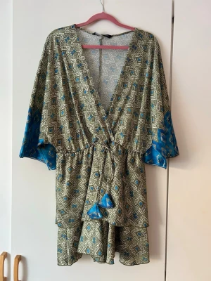 Mönstrad omlottklänning med blå detaljer - Kort omlottklänning med geometriskt mönster i beige, blått och svart. Vida trekvartsärmar med blå kontrastkant och knytband i midjan med tofsar. Klänningen har volangkant nedtill och djup V-ringning. Materialet känns lätt och luftigt.