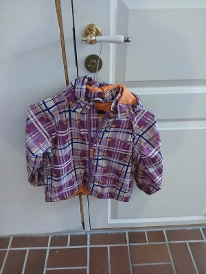 Lila rutmönstrad vindjacka med huva - Cool vindjacka med rutigt mönster i lila, rosa, blått och vitt. Jackan har en huva, dragkedja framtill och orange foder. Perfekt för att hålla vinden borta och ge en färgstark look. Elastiska ärmslut och lätt material gör den smidig att bära. (Står namn i den!!)