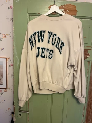 Beige New York Jets sweatshirt - Oversized beige sweatshirt med stort 'NEW YORK JETS'-tryck i grönt och svart på ryggen. Tröjan har rund halsringning, långa ärmar och ribbade muddar vid ärmslut och nederkant. Storlek M. Be gärna om bilder, priset kan alltid diskuteras!