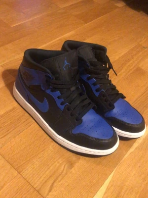 Air Jordan 1 Mid Svart/Blå - Säljer ett par Air Jordan 1 Mid sneakers i svart och blått med vit sula. Klassisk siluett med svart läder och blå detaljer på tå, häl och Swoosh. Skorna har svarta skosnören och ikoniska Jordan-loggor på sidan och plösen. Perfekta för dig som gillar streetwear.