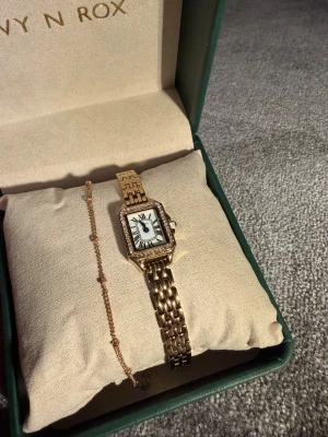 Klocka  - Damklocka, rektangulär i guld. Armbandet kom med när klockan köptes. Länken har kortats, extra biten följer med i påse. 