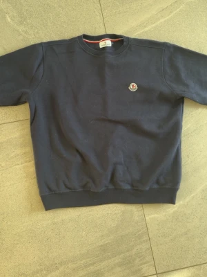 Mörkblå sweatshirt från Moncler - Mörkblå sweatshirt från Moncler med rund halsringning och ribbade muddar vid ärmslut och nederkant. Tröjan har en liten Moncler-logga på bröstet och är tillverkad i mjukt bomullsmaterial. Enkel och stilren design som passar till det mesta.