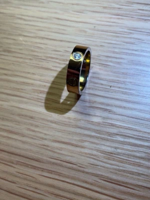 Guldfärgad ring med sten - Snygg guldfärgad ring i metall med en klar sten infattad och dekorativa skruvliknande detaljer runt om. Ringen har en bred, slät design som ger ett modernt och stilrent intryck.