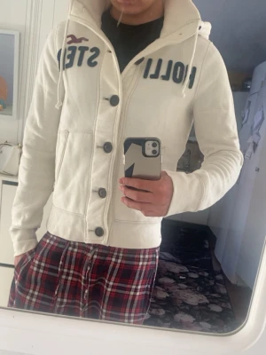 Vit hoodie från Hollister med knappar - Vit hoodie från Hollister med stora knappar framtill, dragsko i huvan och broderad text på bröstet. Klassisk college-stil med fickor på sidorna och ribbade muddar. Perfekt för en avslappnad look och enkel att matcha med jeans eller mjukisbyxor.