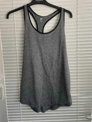 Grått sportlinne från H&M Sport - Snyggt grått sportlinne från H&M Sport med svarta kantband och brottarrygg. Tillverkat i ett mjukt och stretchigt funktionsmaterial som andas, perfekt för träning och gym. Enkel och stilren design som passar till alla typer av sportaktiviteter.